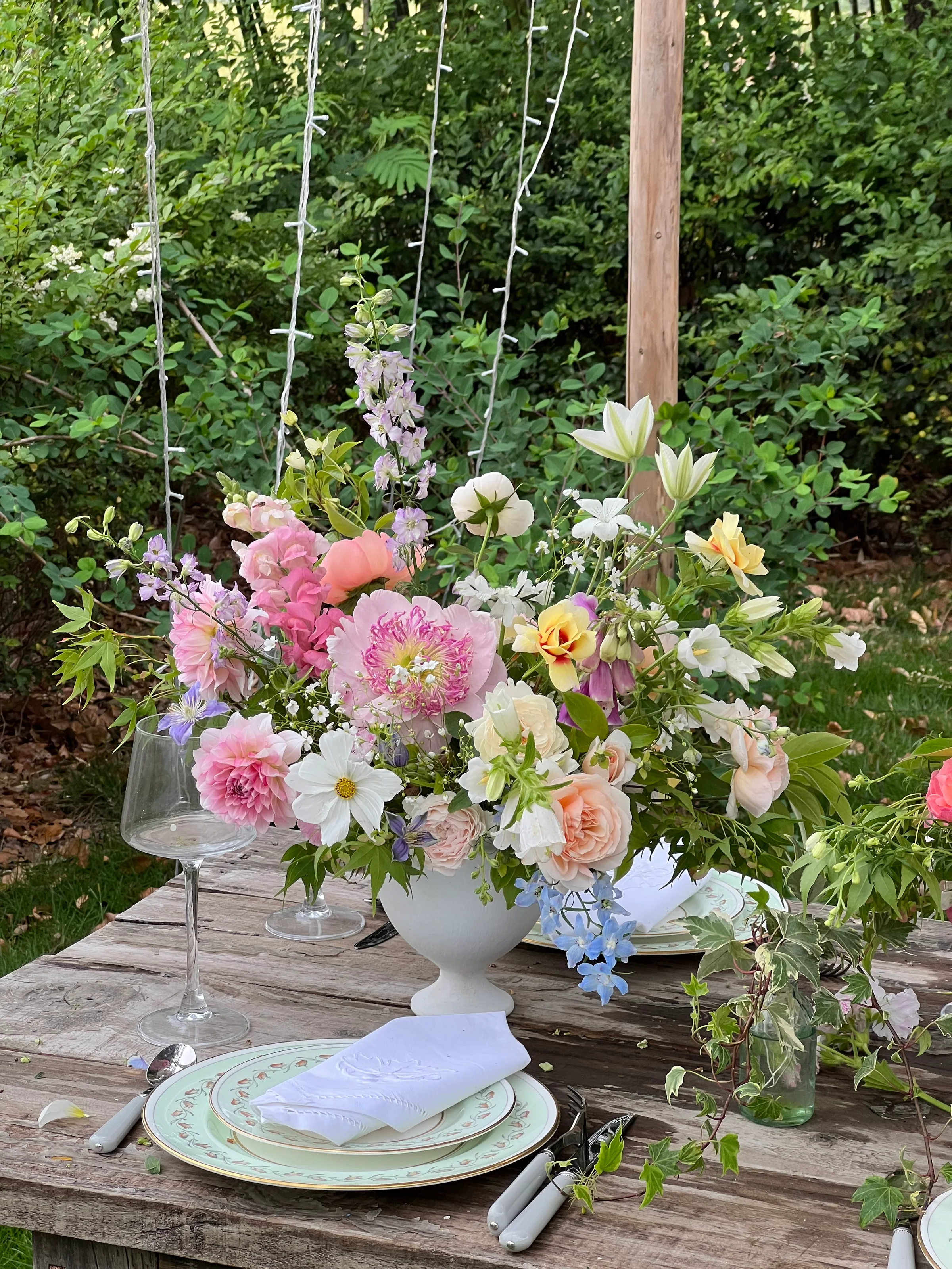 Pastel garden centerpiece of peonies, garden roses, and cosmos in a white footed vase on a rustic outdoor table