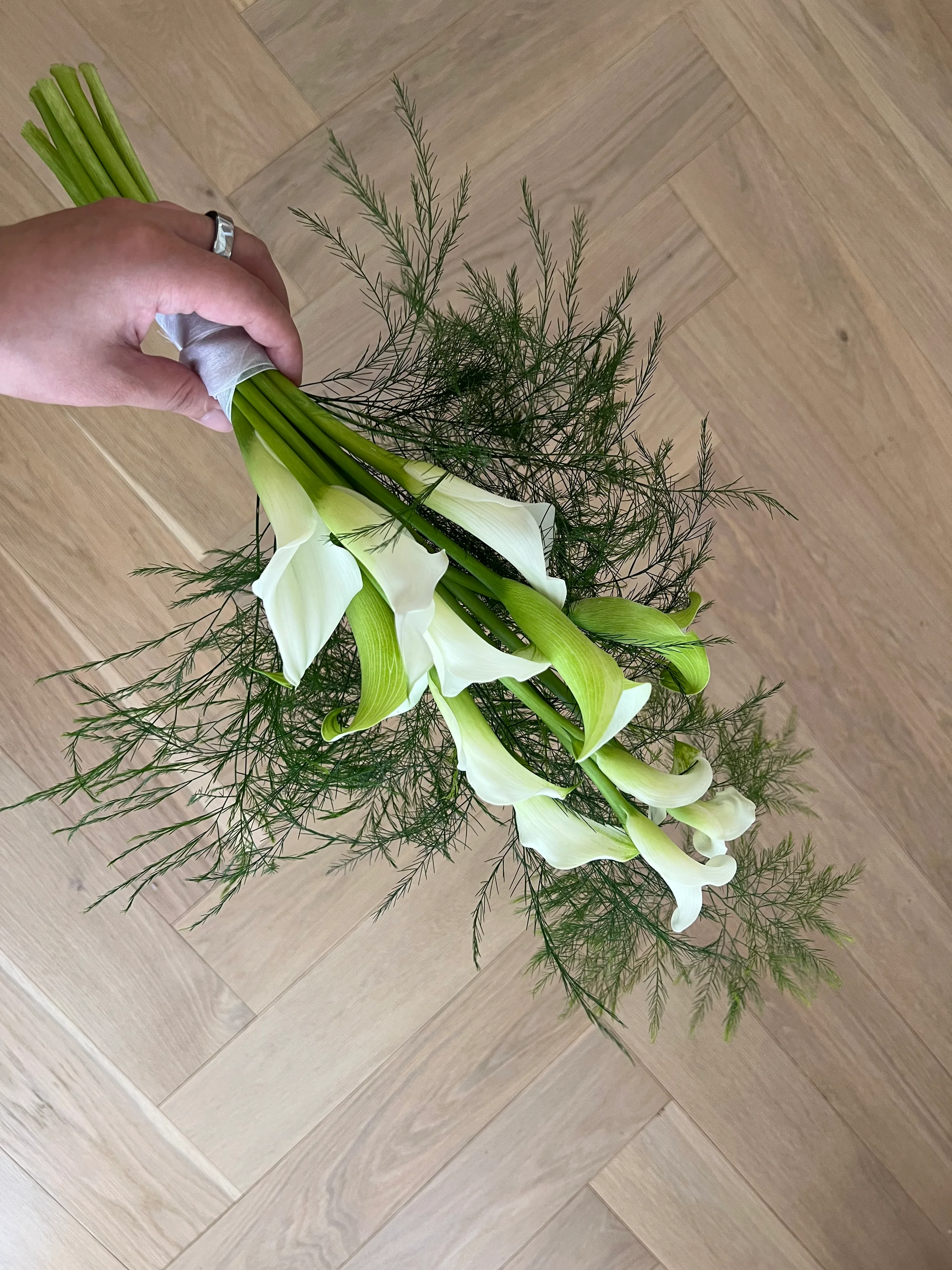 Hand holding a white calla lily bouquet with feathery greenery over a herringbone wood floor