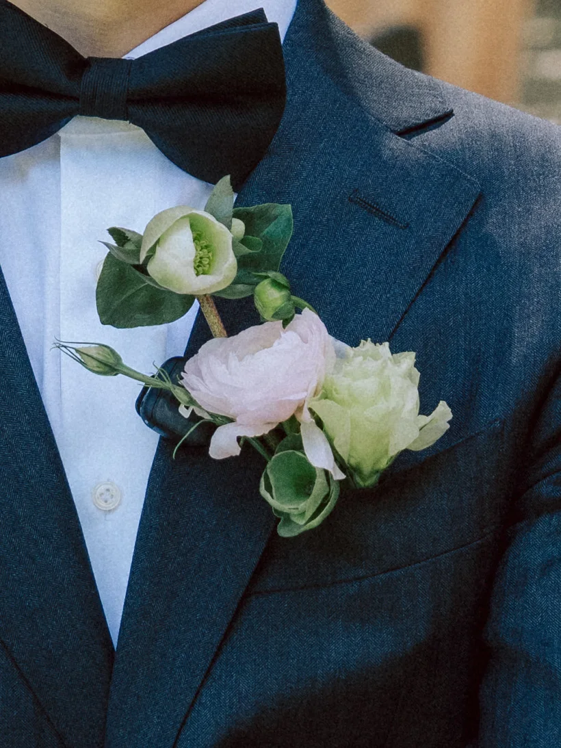 White and blush boutonniere with green buds pinned to a dark suit lapel beneath a black bow tie