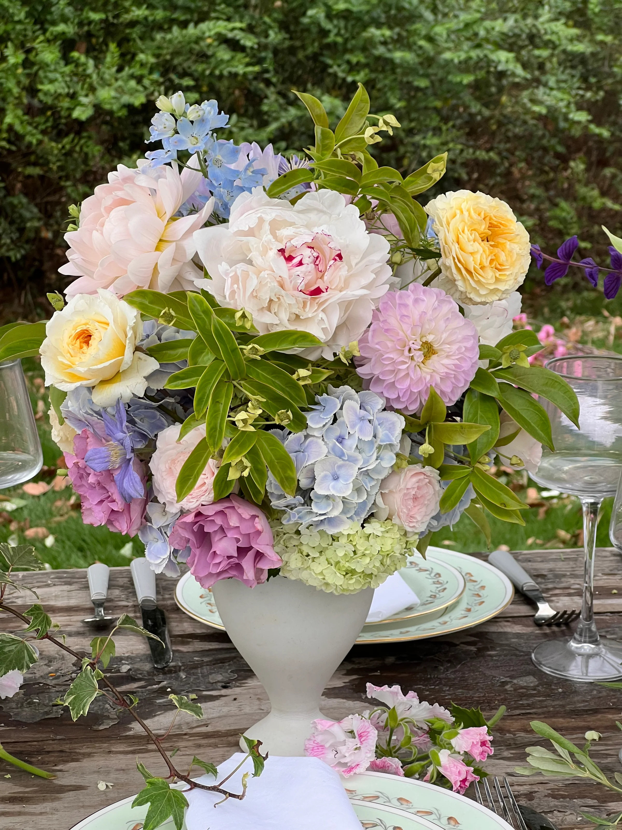 Pink and yellow roses with pale blue hydrangeas in a white footed vase on an outdoor table
