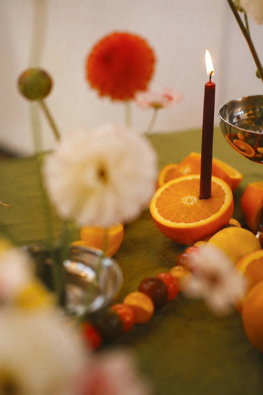 A lit burgundy taper candle in an orange half framed by softly blurred white flowers and an orange dahlia