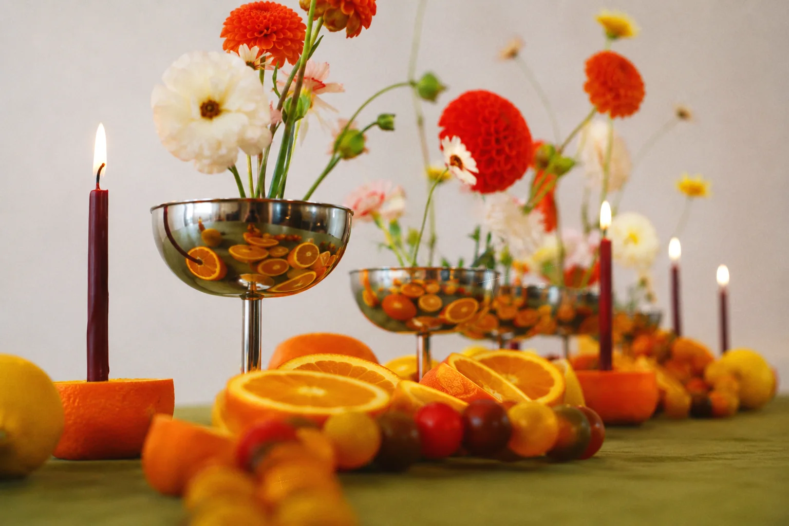 Low-angle view along a citrus tablescape with silver floral vessels, orange dahlias, pale blooms, and lit taper candles