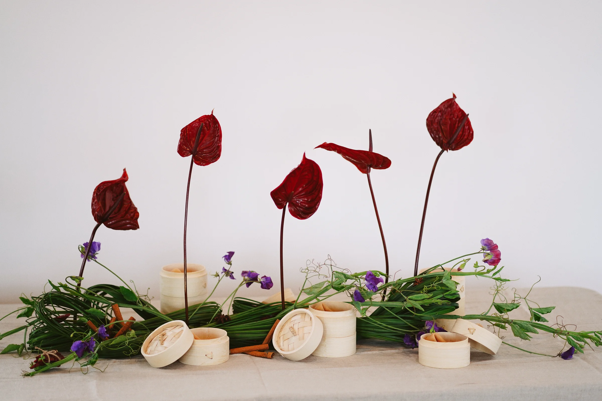 Sculptural burgundy anthuriums on slender stems above a low arrangement of looping leaves, purple flowers, and bamboo steamers
