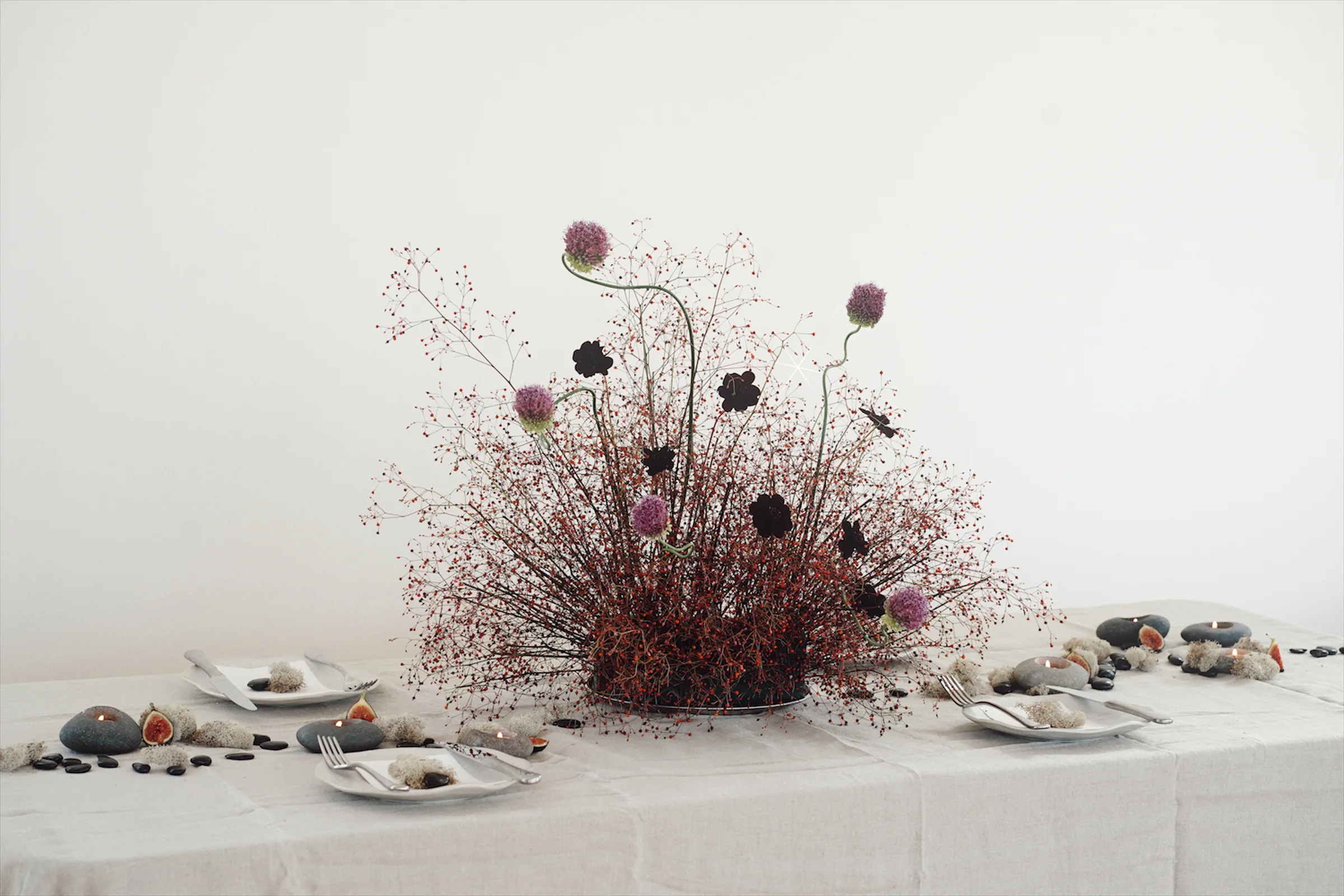 Wide editorial tablescape with a sculptural floral arrangement of red berry stems, purple alliums, dark blooms, stone candles, figs, and minimal place settings