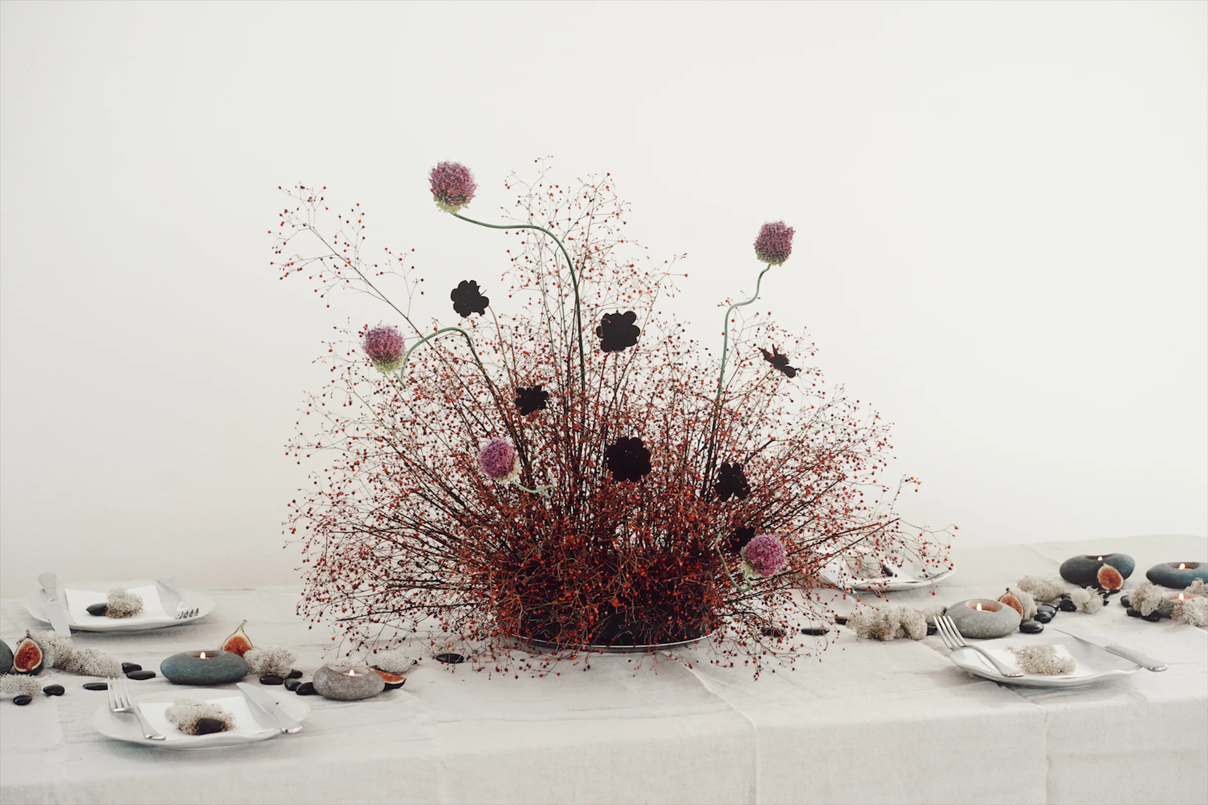 Centered sculptural floral arrangement with red berry stems, purple alliums, dark flowers, figs, stone candles, and white place settings