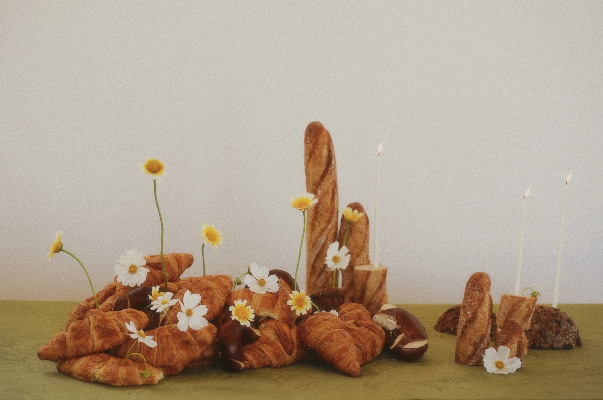 Wide tablescape of croissants, upright baguettes, and rustic loaves interspersed with white and yellow flowers and slender candles