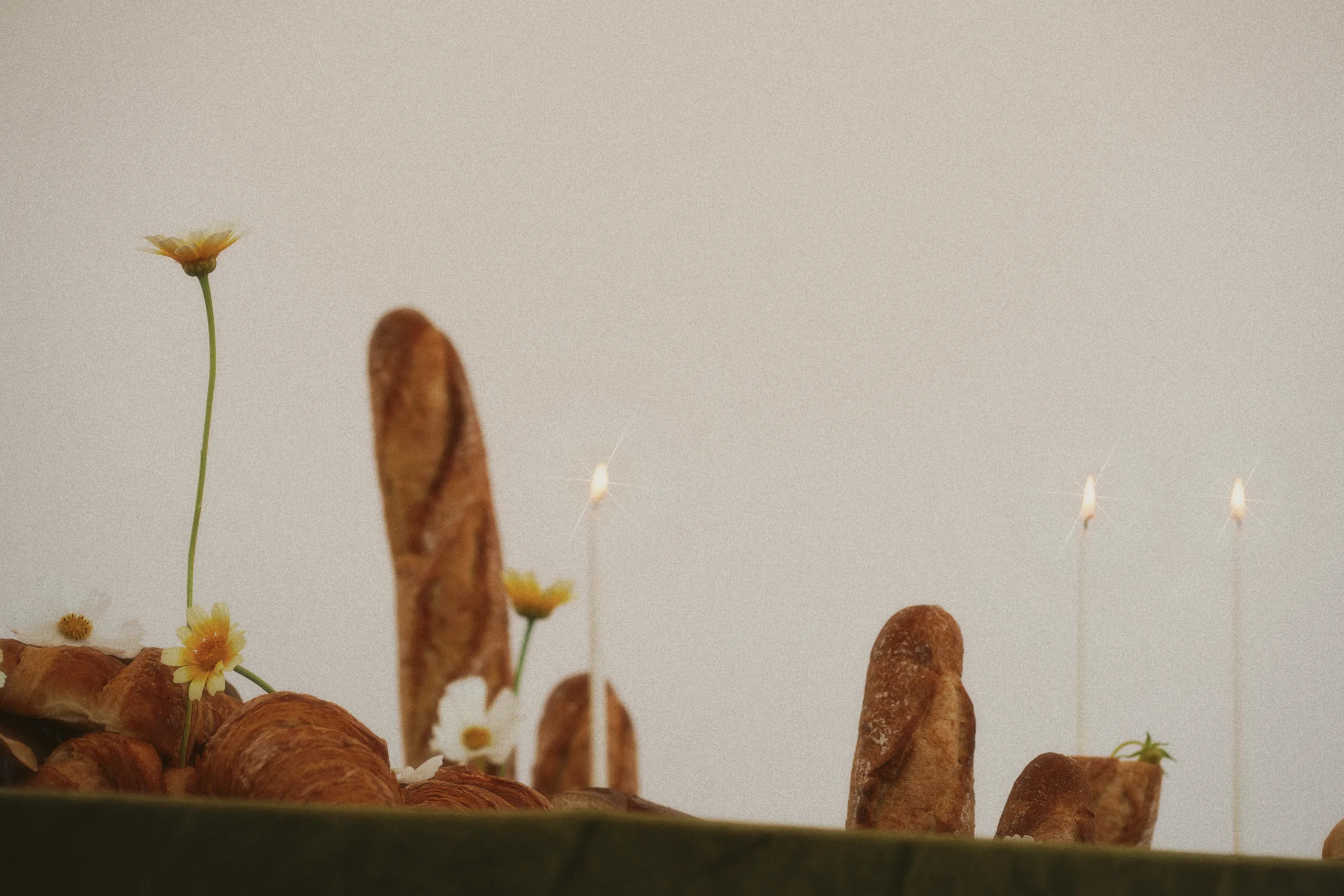 Low-angle view of upright baguettes, delicate yellow flowers, and lit white taper candles against a neutral backdrop