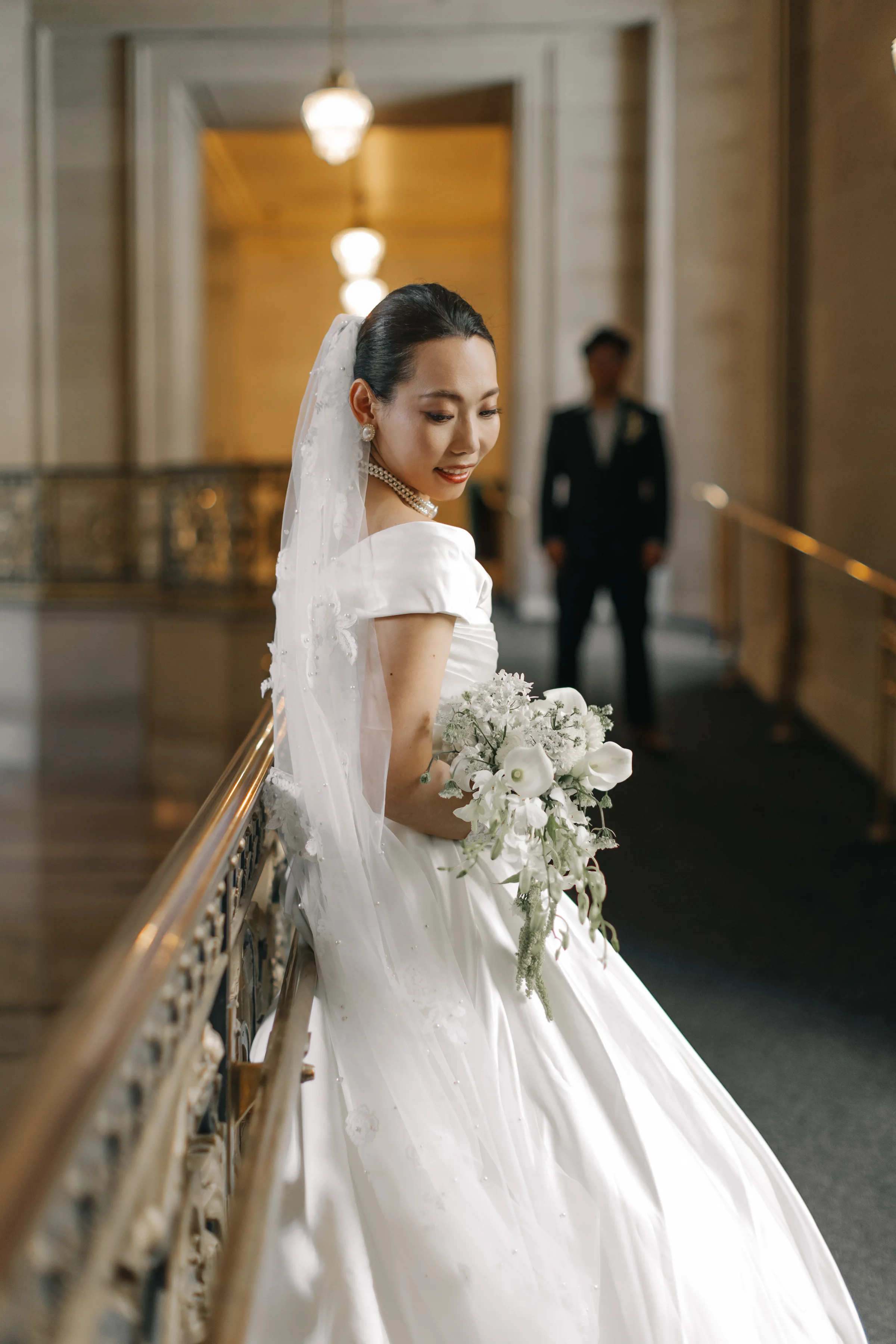 Bride at the City Hall balustrade holding a white bouquet, the groom blurred behind