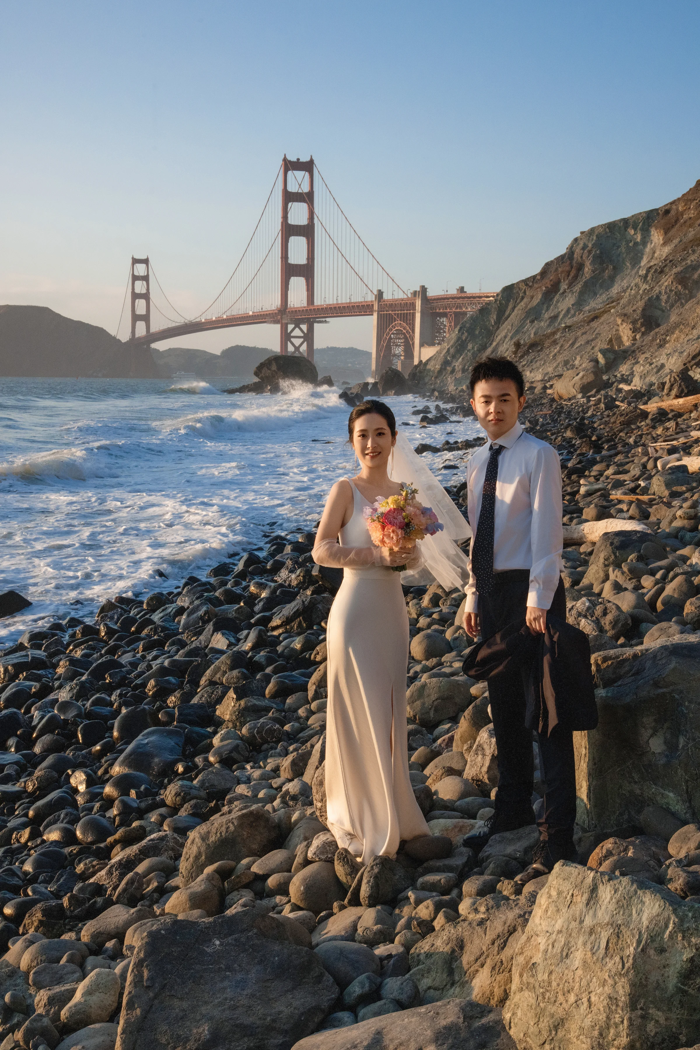 Couple by the rocky shore at sunset with the Golden Gate Bridge behind, the bride holding a bouquet