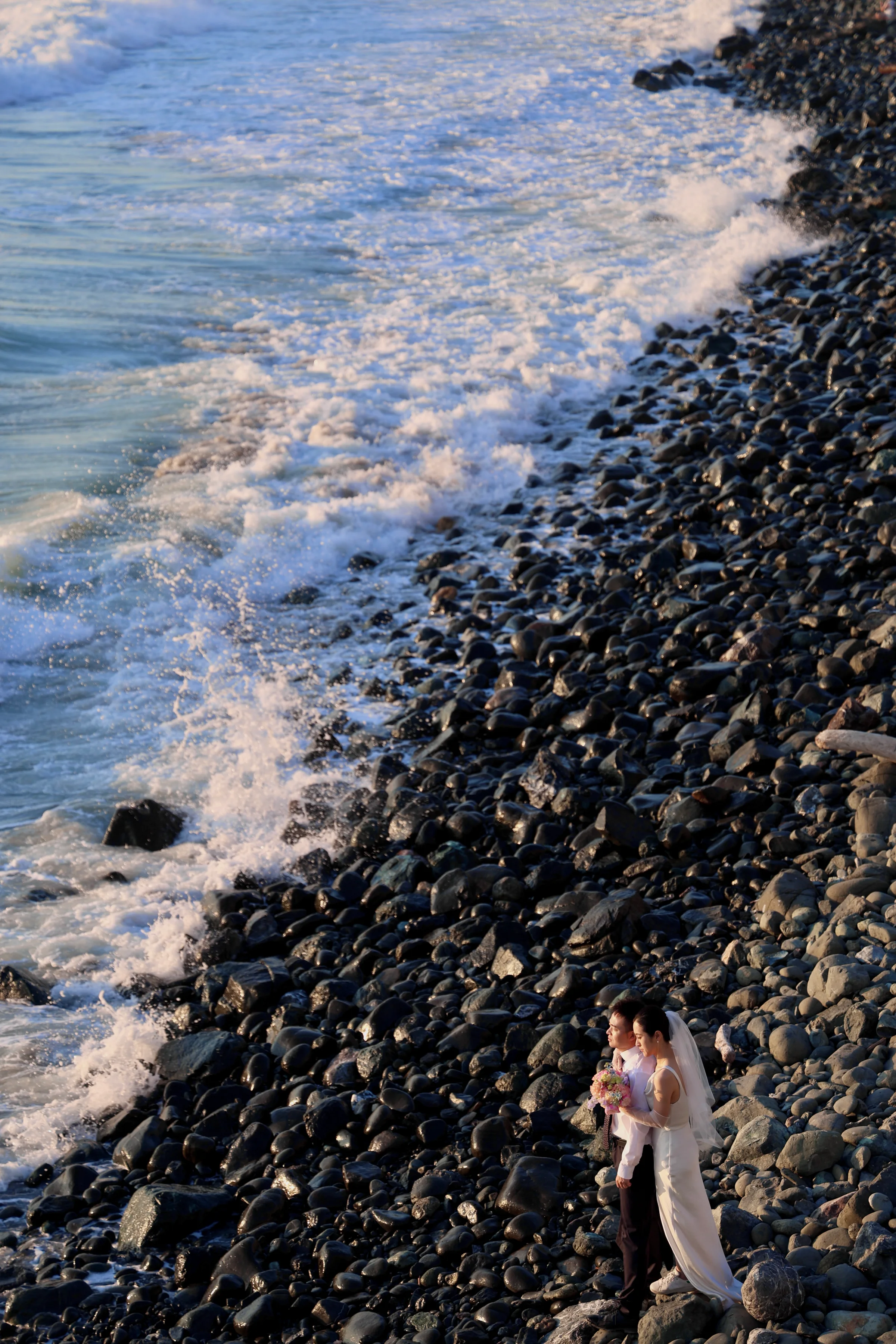 Bride in white small against a rocky, surf-lined beach