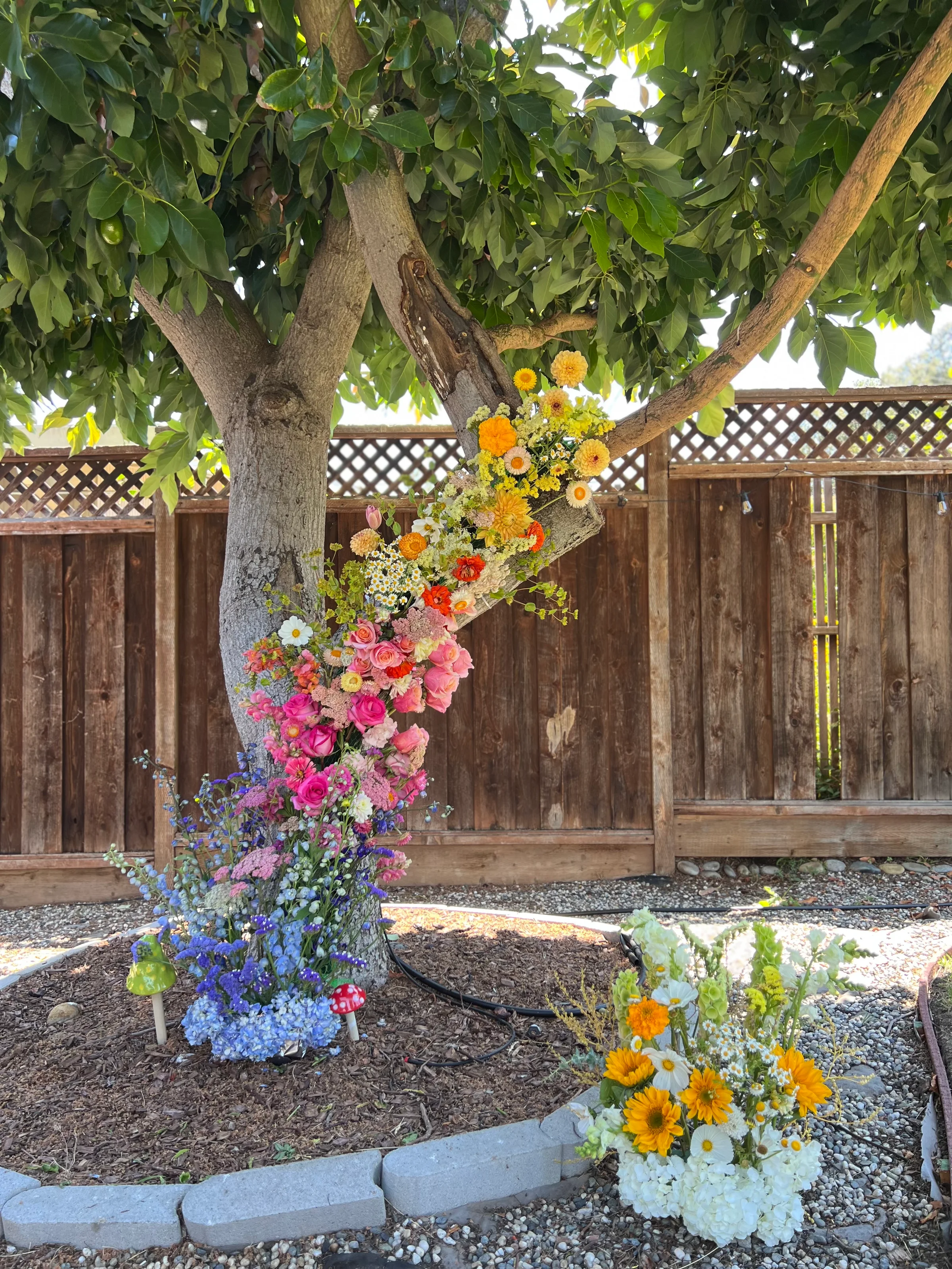 Cascading pink, yellow, and blue floral installation climbing a backyard tree with ground arrangements