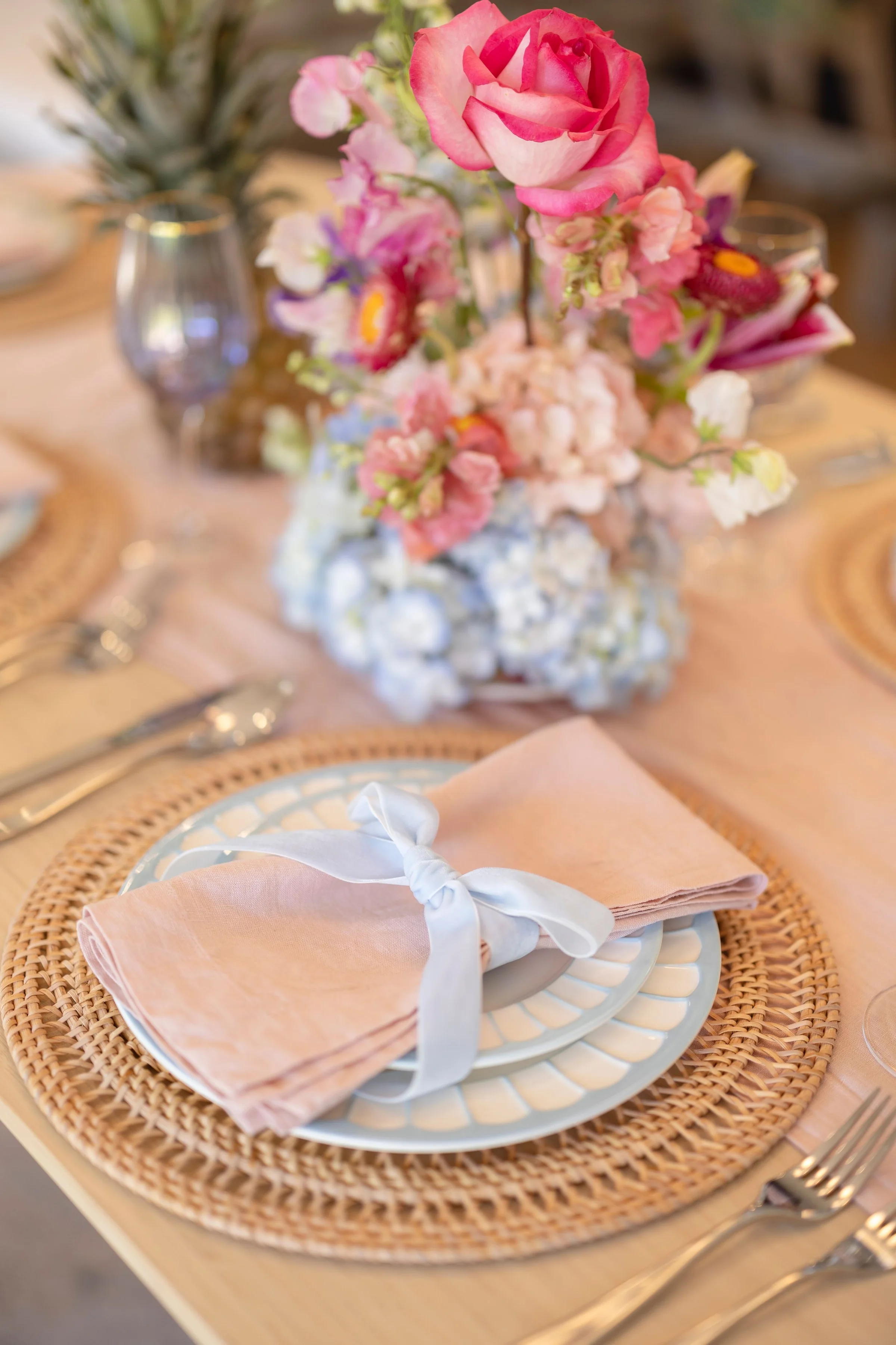 Place setting with a blush napkin and blue velvet bow on a rattan charger, pink and blue centerpiece behind