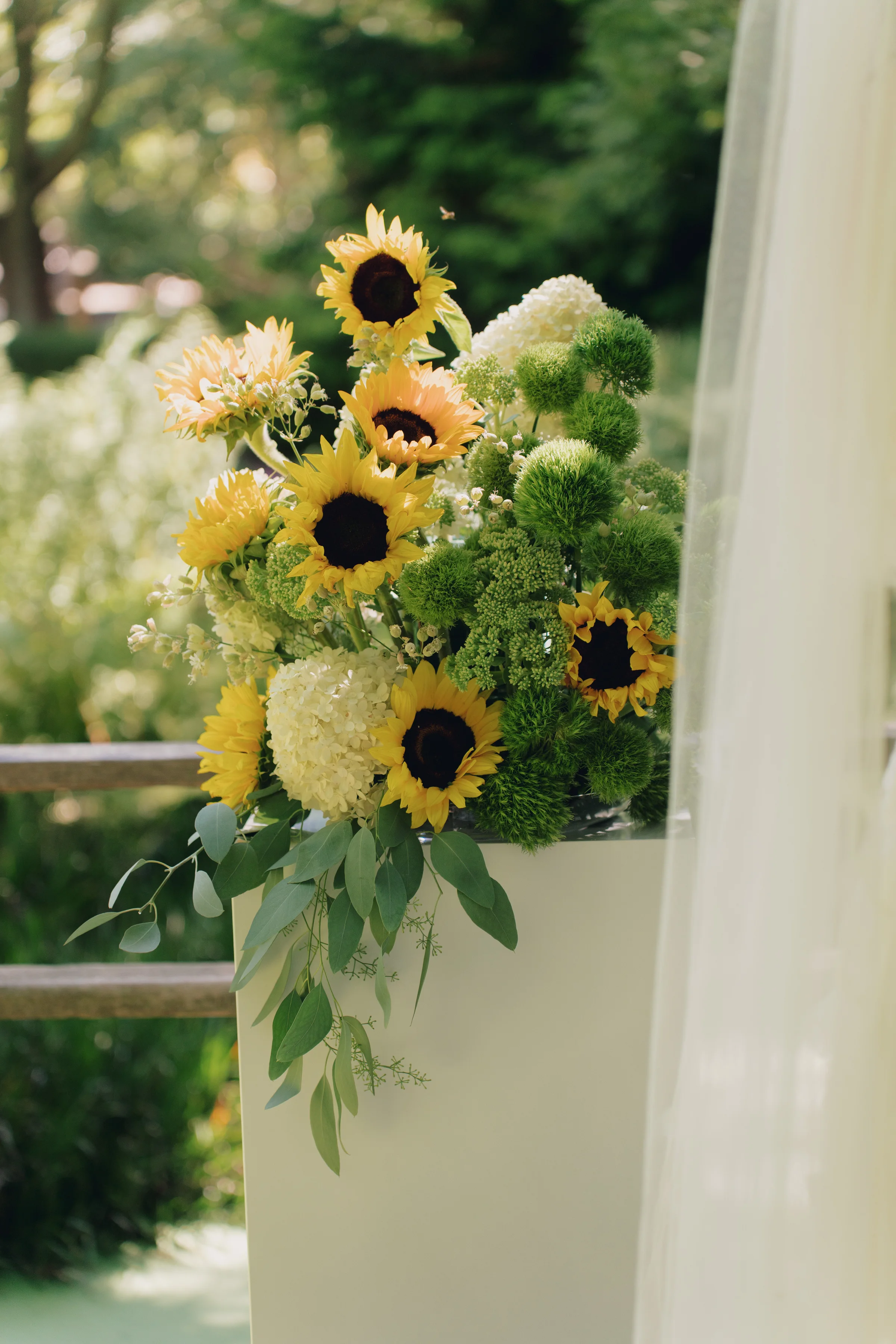 Ceremony arrangement of sunflowers, white hydrangeas, green blooms, and cascading eucalyptus on a white pedestal.