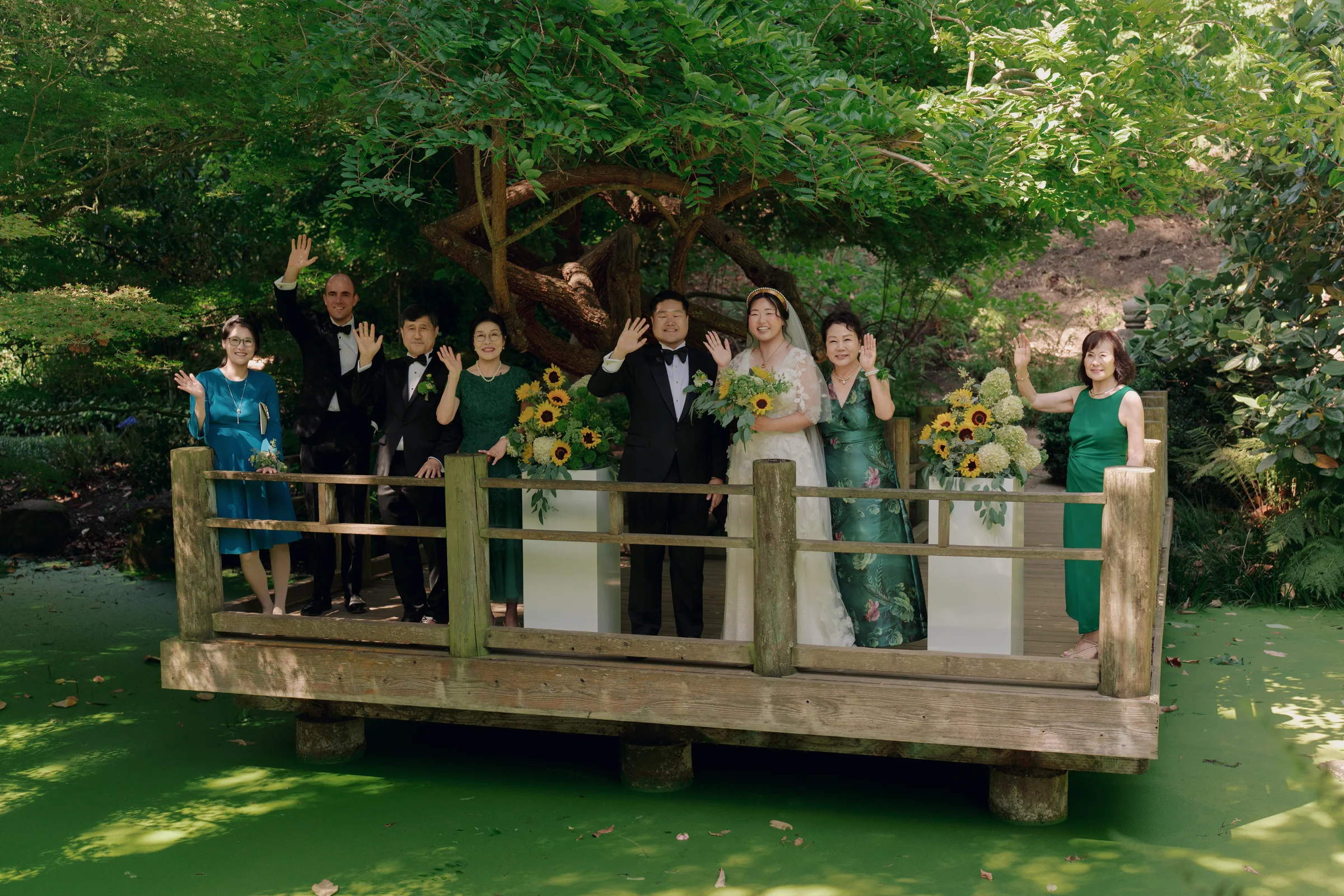 Newlyweds and family wave from a wooden garden bridge framed by sunflower floral arrangements.