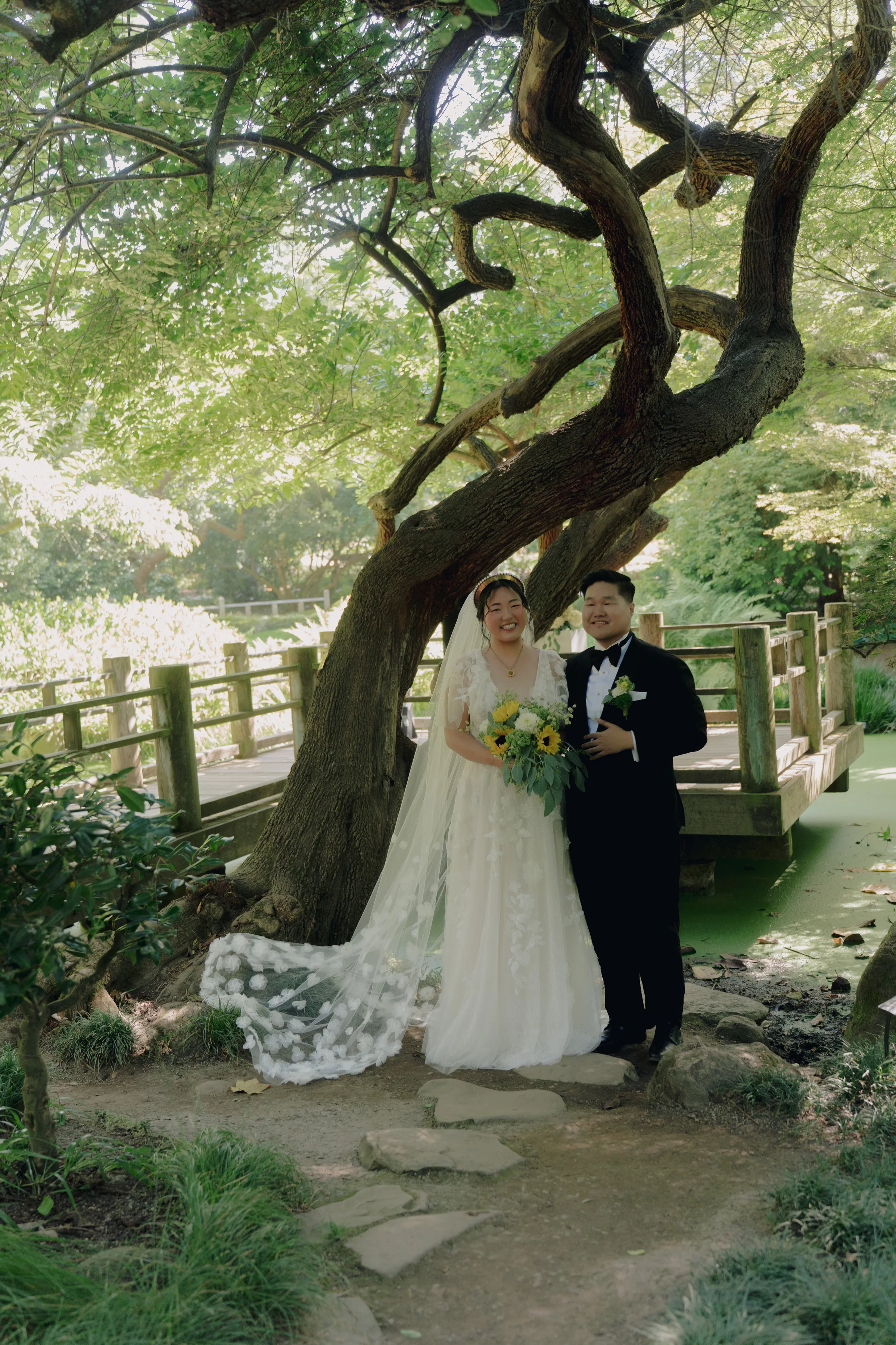 Newlyweds pose beneath a sculptural tree beside a wooden bridge in the Moon Viewing Garden.