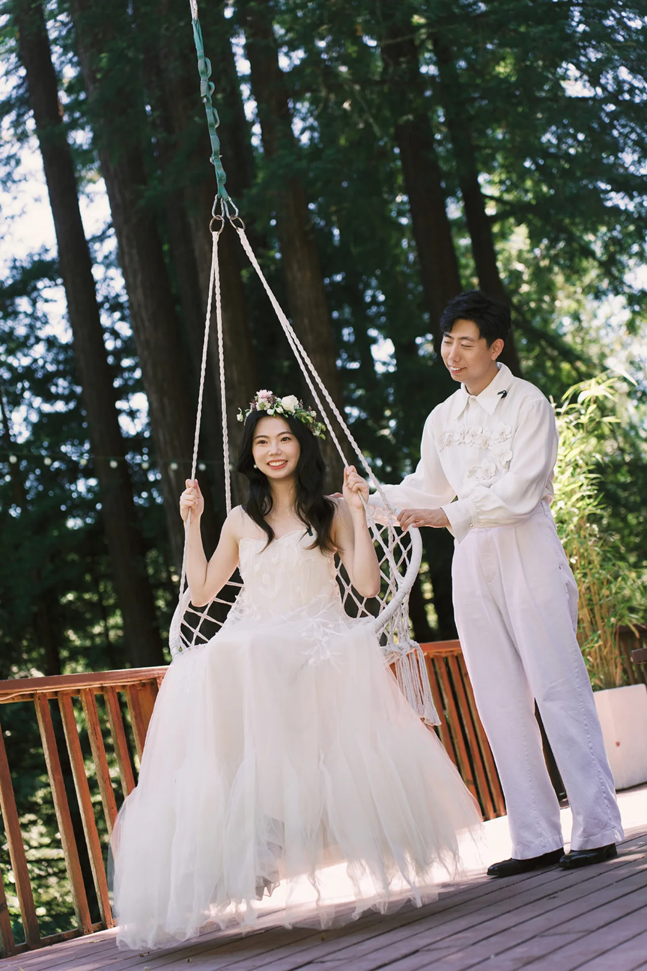 Bride in a floral crown seated on a hanging chair while the groom stands beside her on a redwood deck.