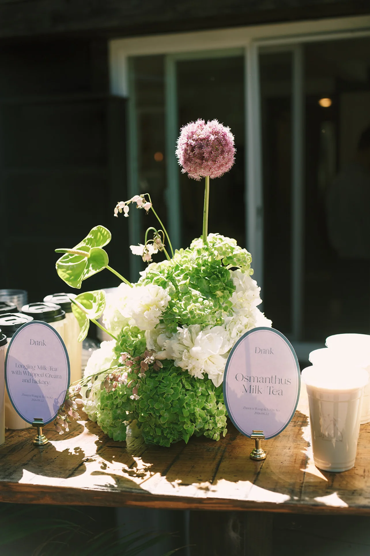 Green and white hydrangeas, anthuriums, and a tall purple allium arranged beside cups of milk tea.