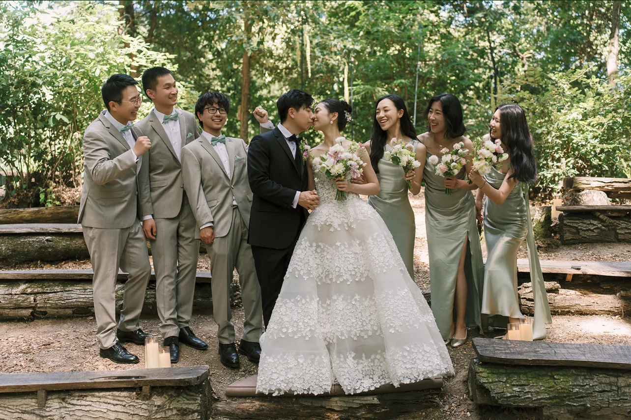 Newlyweds kissing as bridesmaids hold pastel bouquets beside groomsmen in a sunlit redwood grove.
