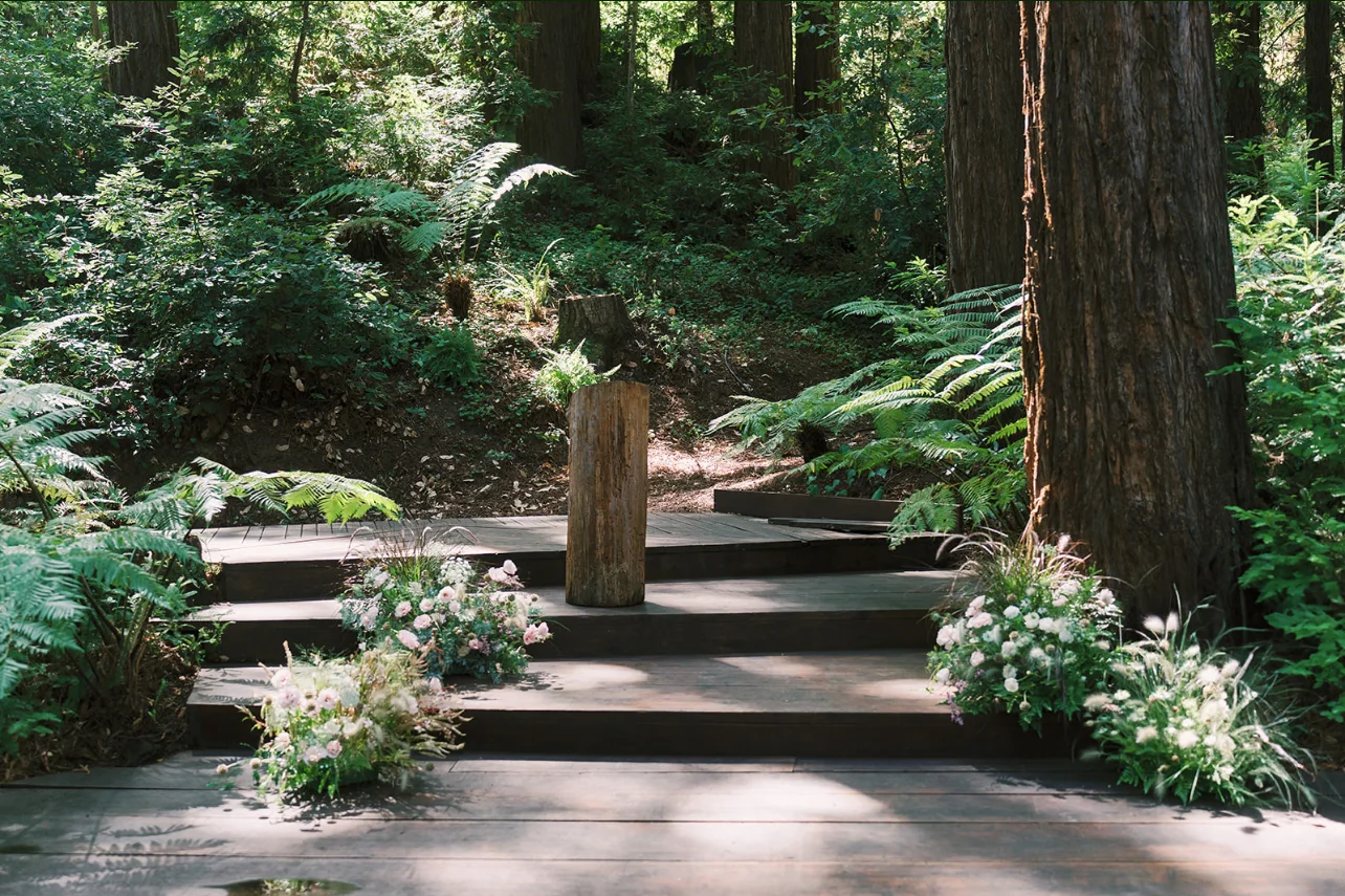 Woodland ceremony platform framed by ferns and low arrangements of pink and white flowers.