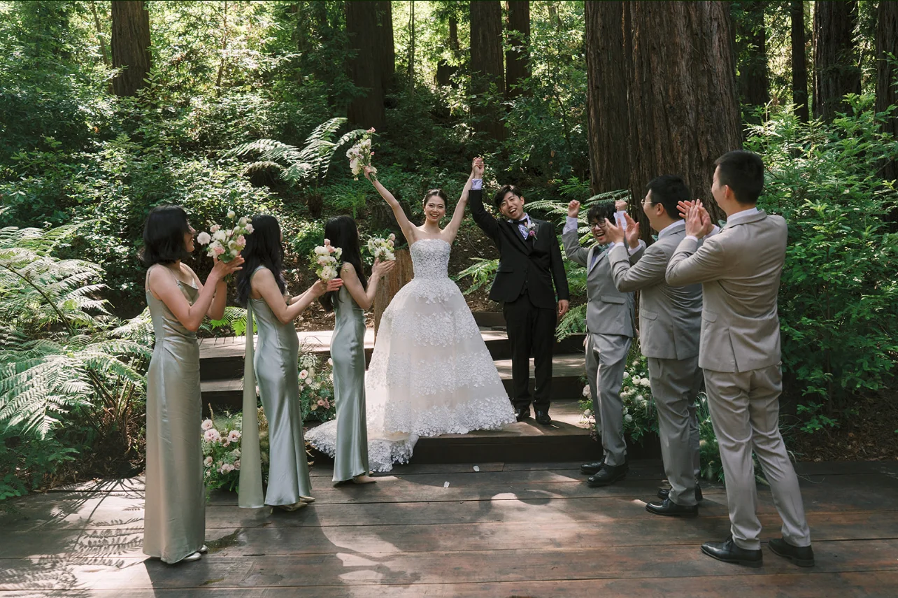 Newlyweds raising their joined hands as attendants cheer beside the woodland ceremony flowers.