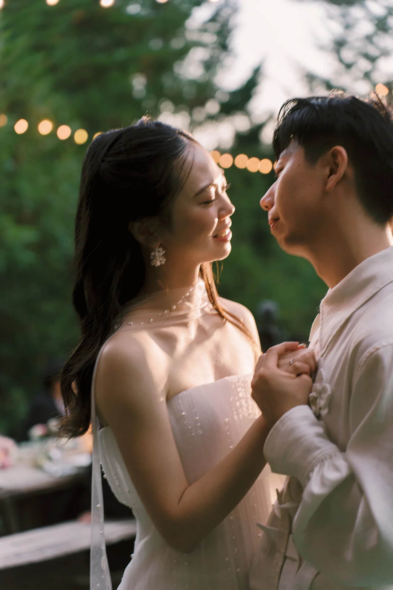 Newlyweds holding hands beneath warm string lights during an outdoor evening portrait.