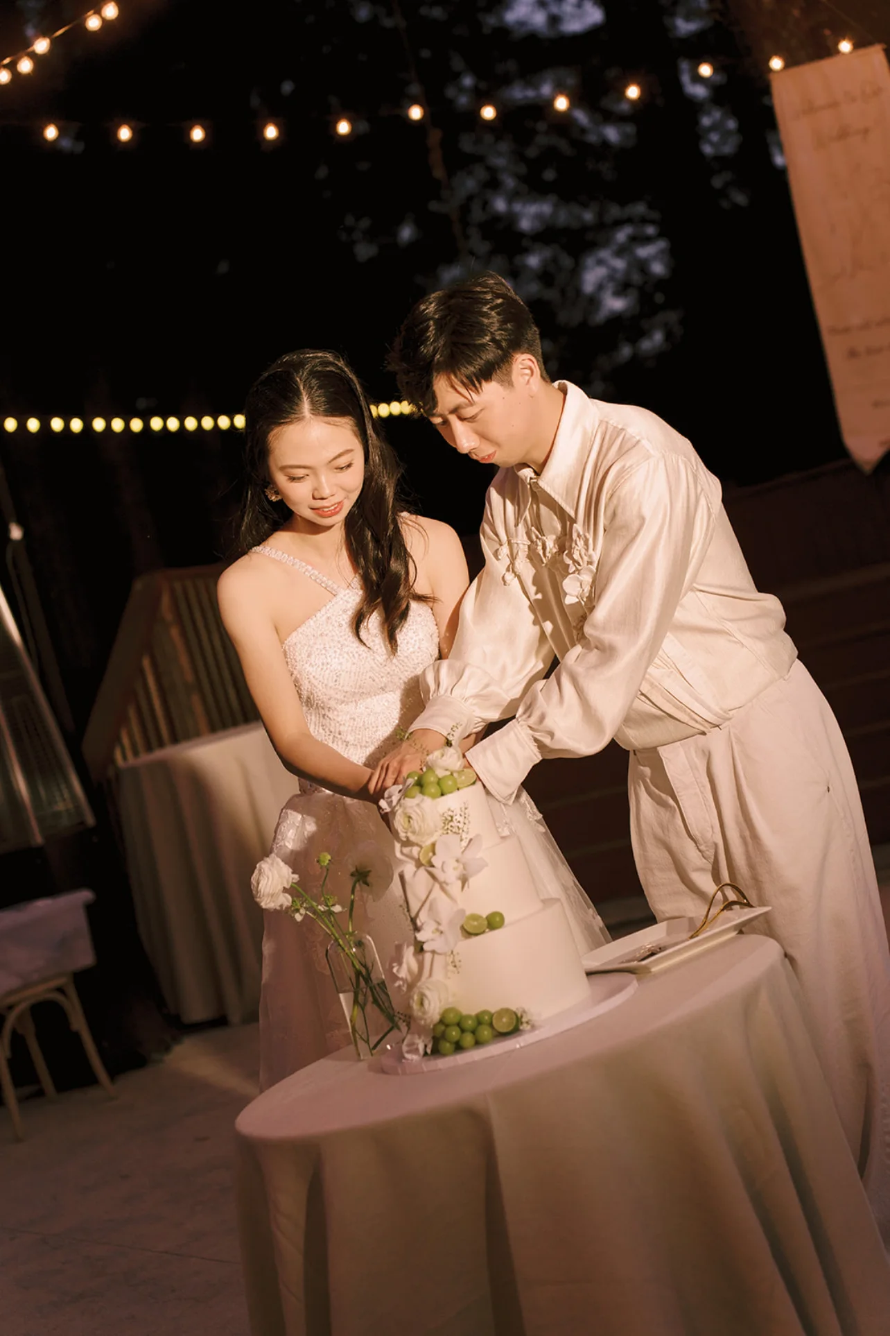 Newlyweds cutting a white wedding cake decorated with white flowers and green grapes beneath string lights.