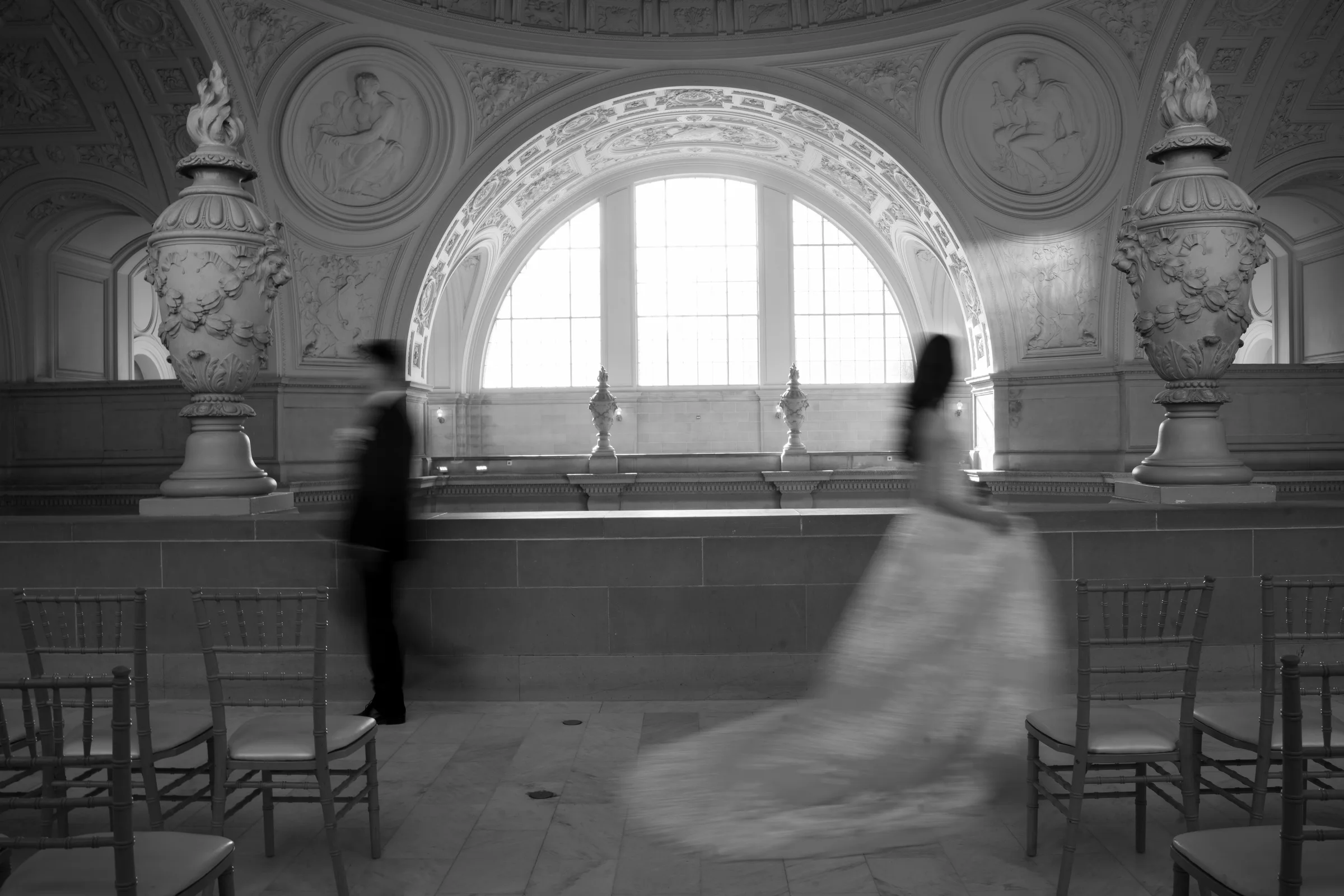 Black-and-white blurred couple walking past rows of chairs beneath the arched window