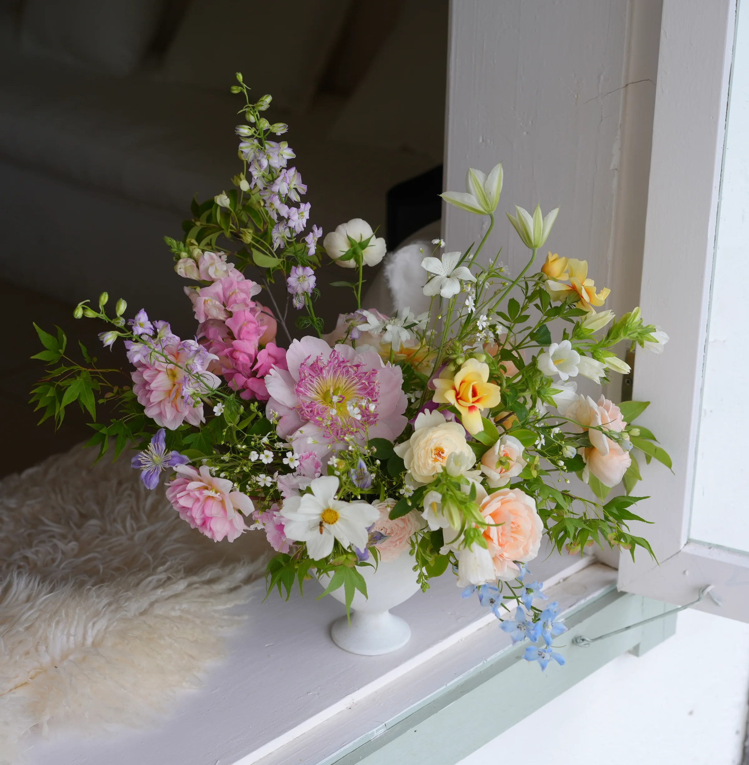 Pastel garden arrangement of peonies, roses, delphinium, and cosmos in a white footed vase on a sunlit windowsill