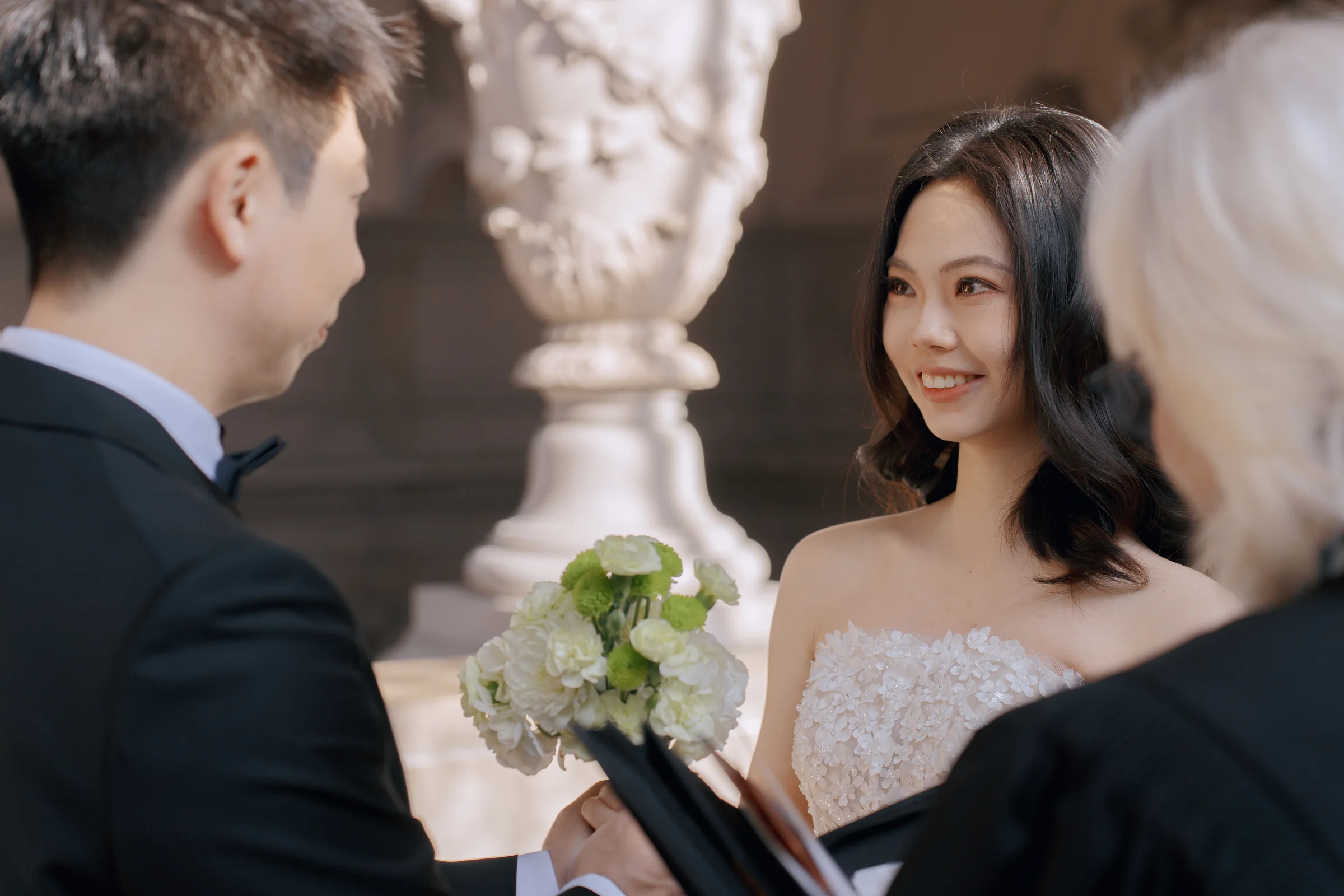 Bride smiling at her partner during a City Hall vow exchange, holding a white bouquet