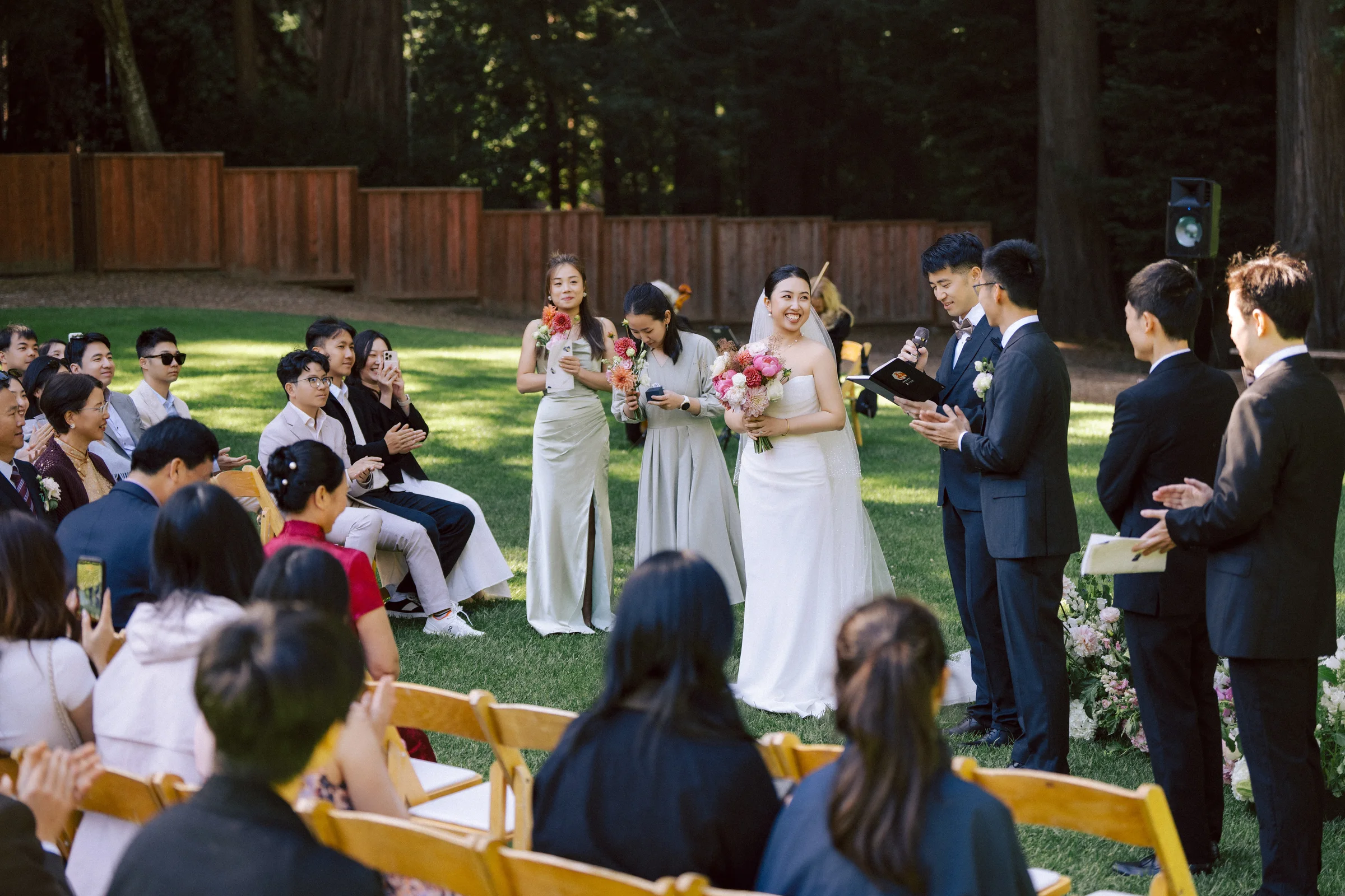 Bride walking a redwood-grove aisle with her bouquet as guests applaud