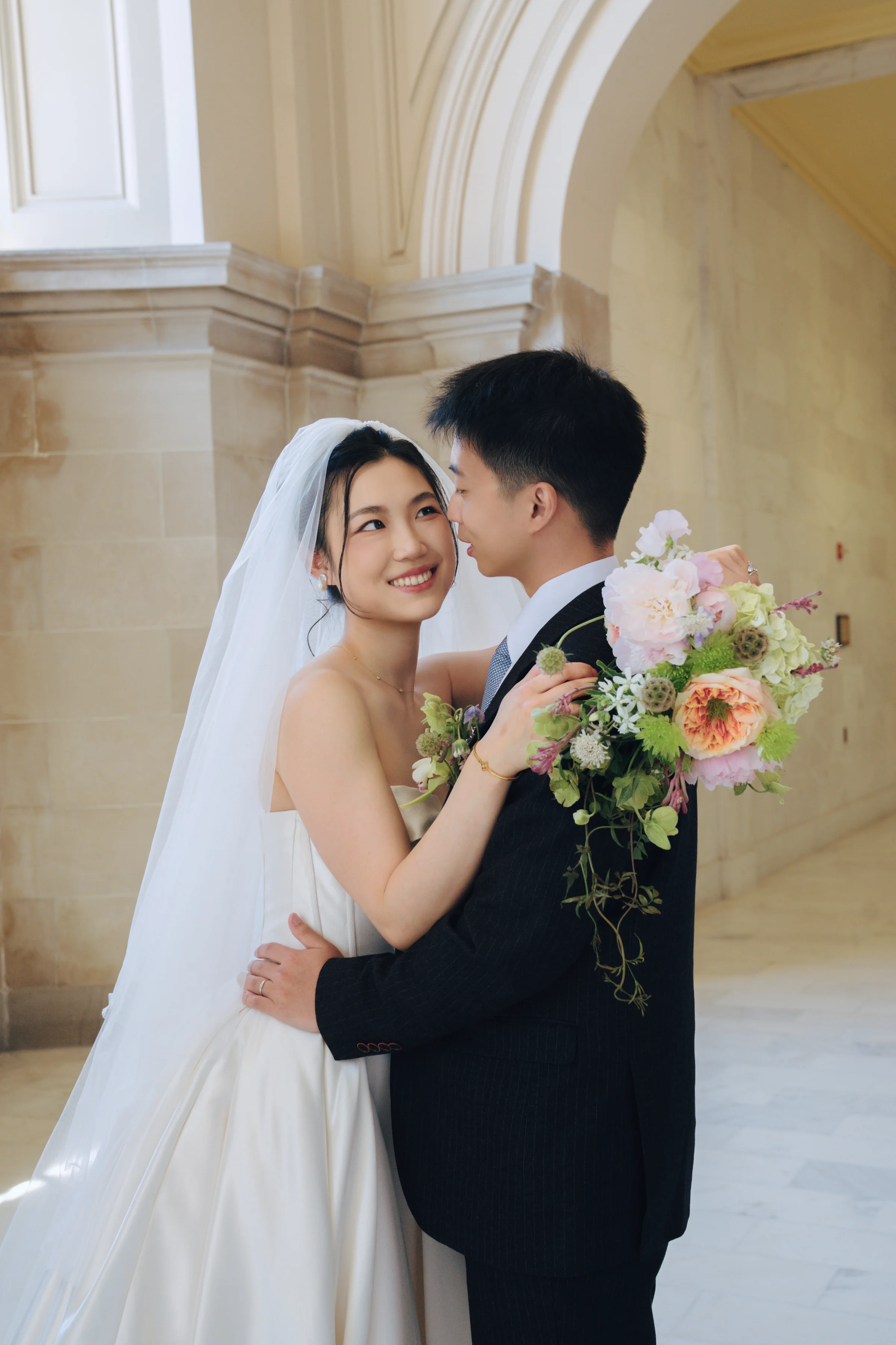 Bride and groom sharing a close moment in a sunlit City Hall archway, the bride holding a pastel garden bouquet