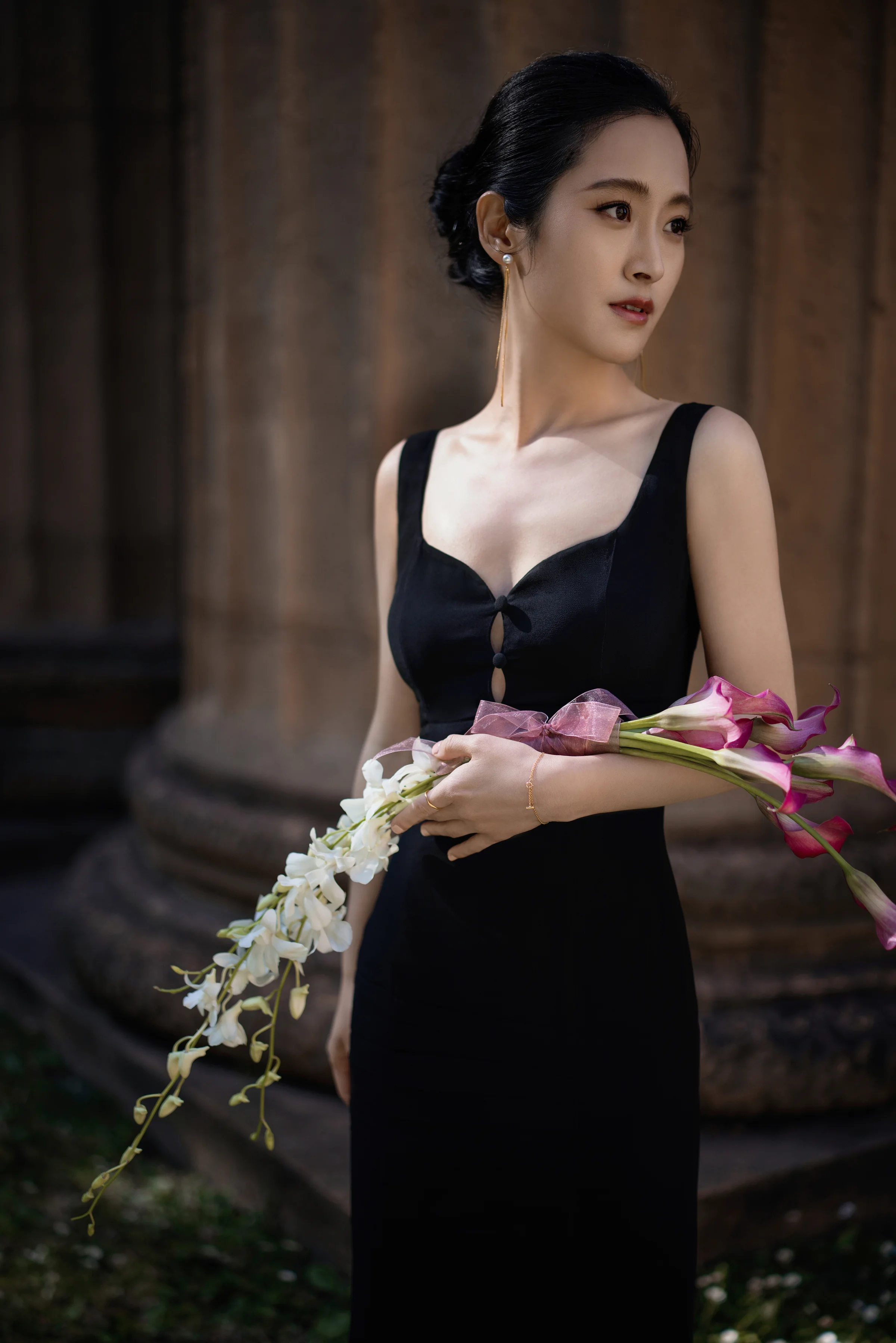 Portrait of a woman in a black gown holding pink calla lilies and trailing white orchids beside a stone column