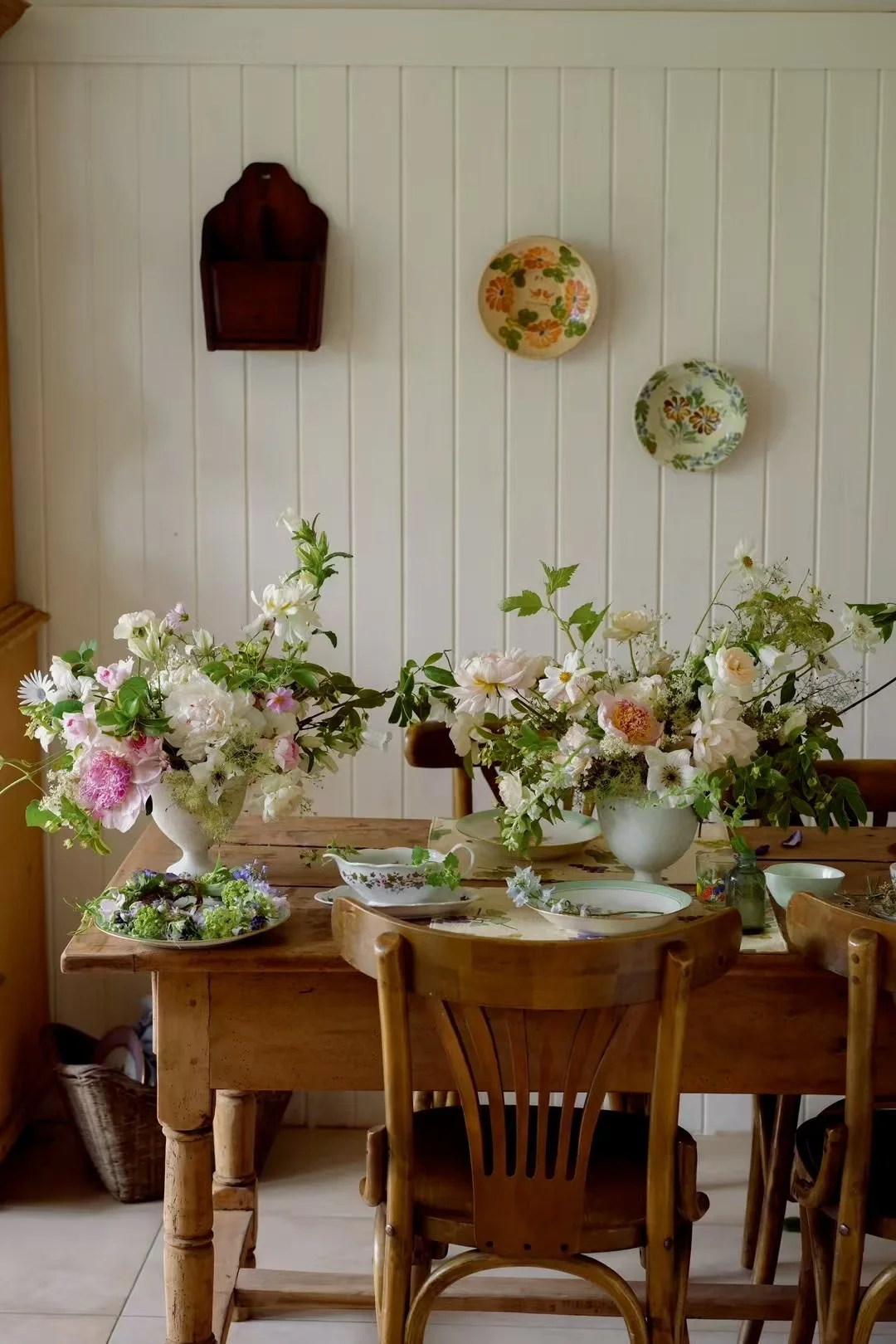Two pastel garden arrangements in white footed vases on a rustic farmhouse table in a cottage dining room