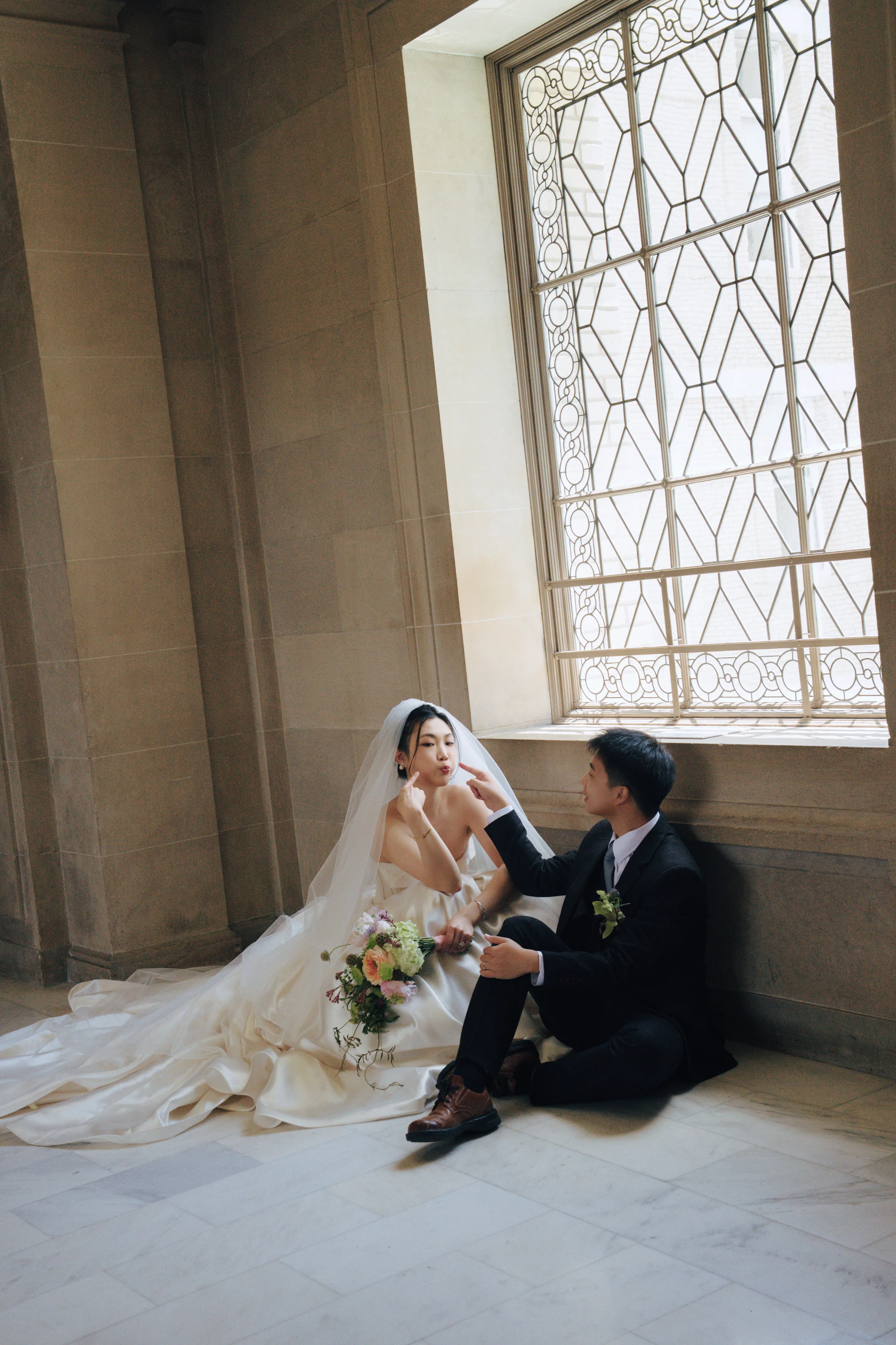 Bride and groom seated by a tall latticed City Hall window, the bride holding a pastel bouquet