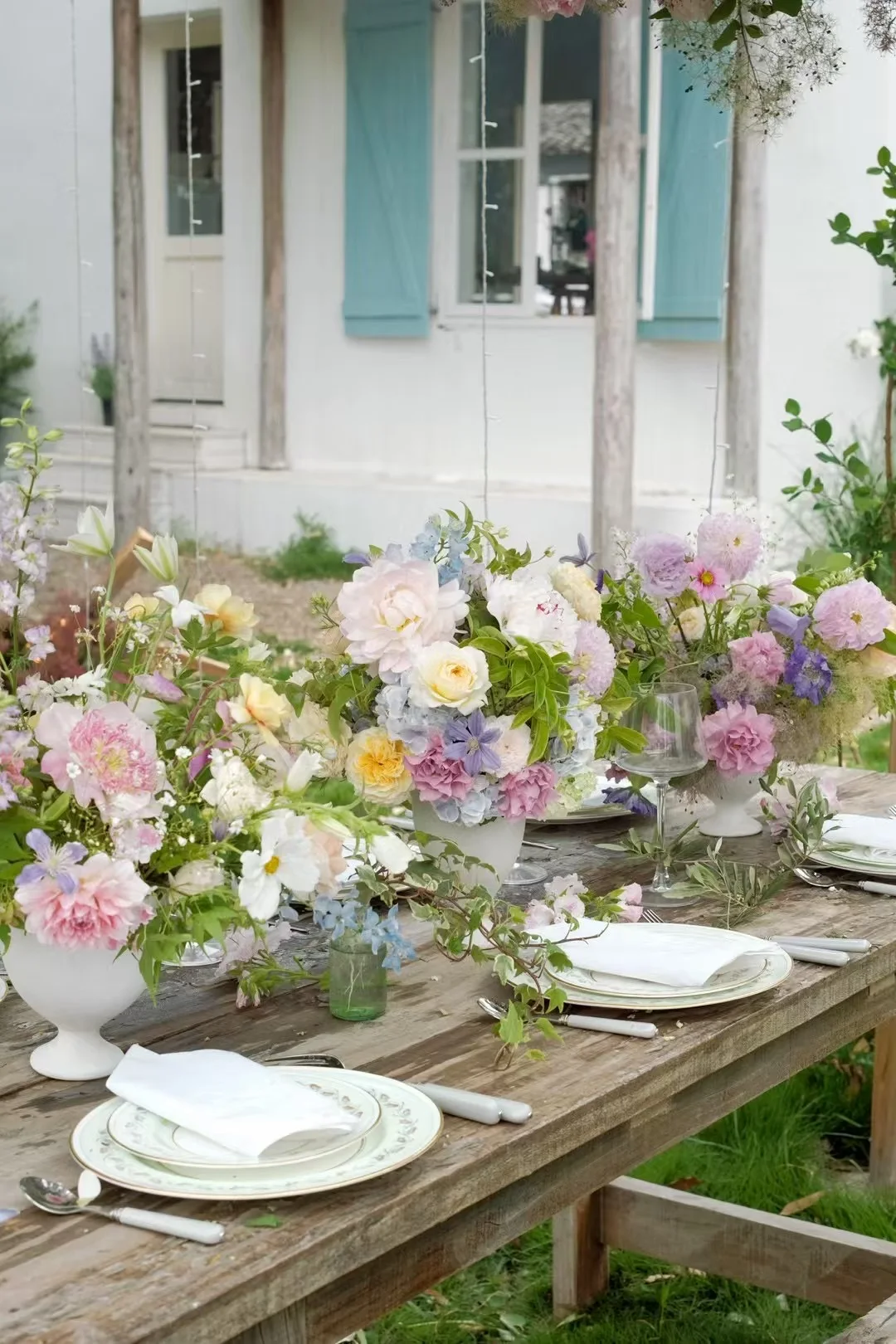 Rustic garden reception table lined with pastel centerpieces before a shuttered cottage